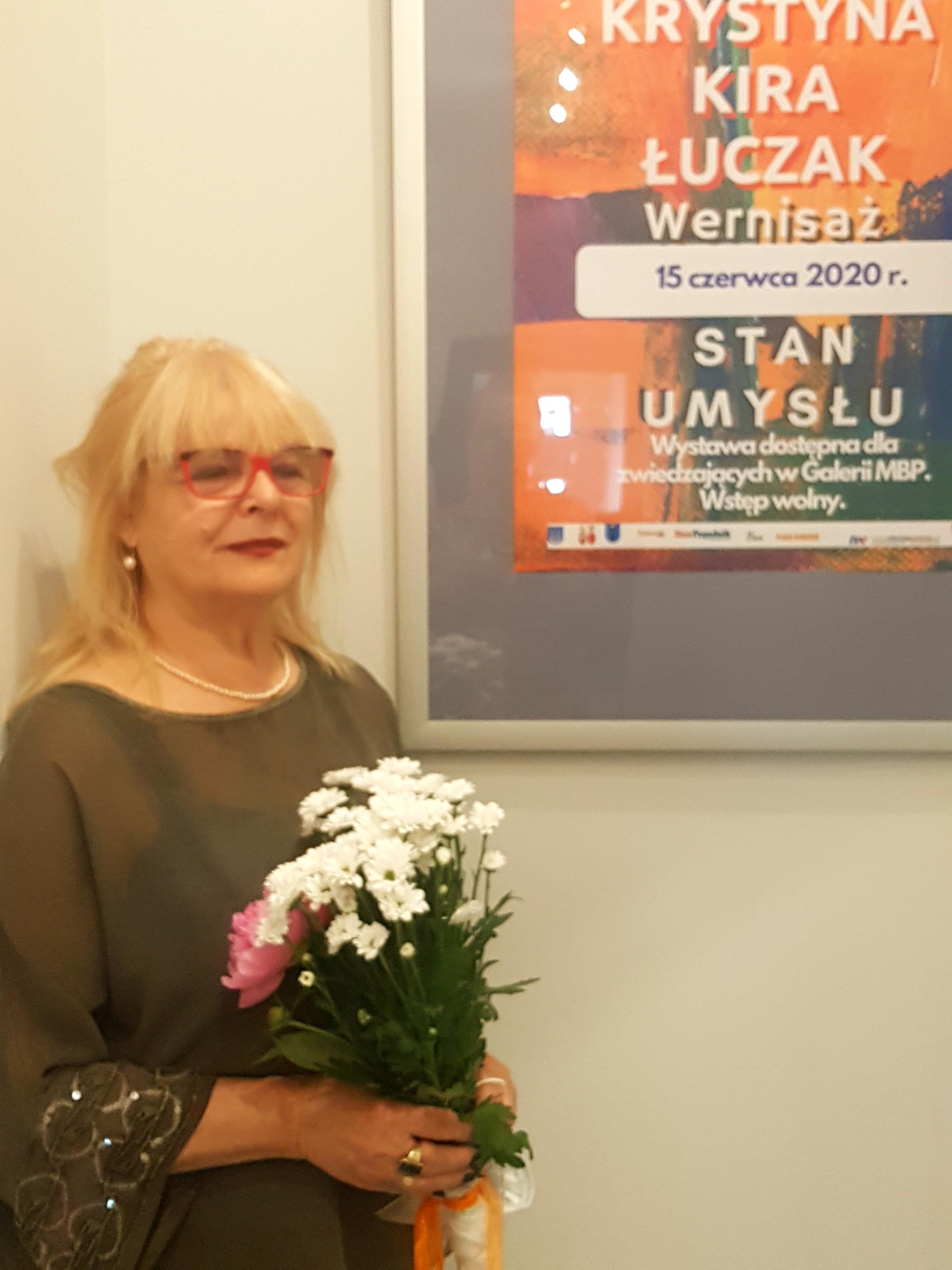 Krystyna Kira Łuczak na tle plakatu zapraszającego na wystawę prac pt. Stan umysłu