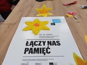 Na stole leży plakat upamiętniający powstanie w getcie warszawskim. Na plakacie widnieje ręcznie wykonany żonkil a niżej jest napis Łączy nas pamięć.