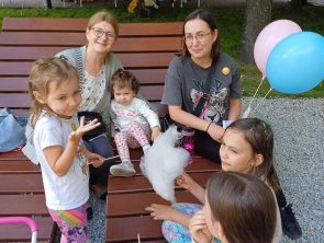 Uczestnicy festynu siedzą na leżakach i dzieci trzymają balony i watę cukrową.