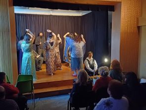 Aktorzy na scenie z podniesioną ręką oraz w głębi aktor z dwoma kijami.