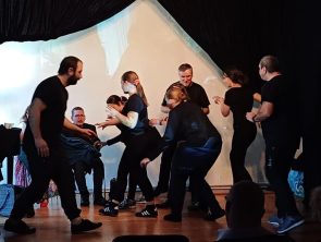 Aktorzy ubrani na czarno poruszają się na scenie.