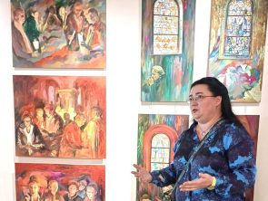 Marzena Sroczyńska na tle swoich obrazów, przedstawiających sceny kościelne