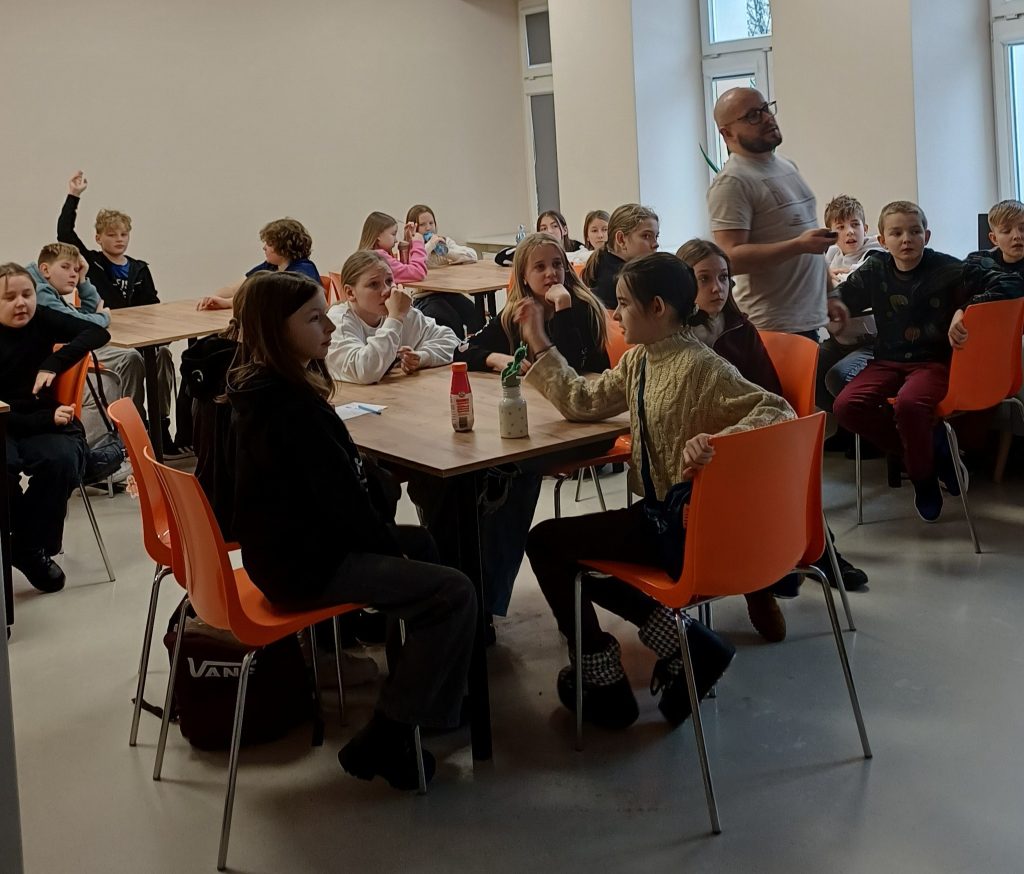 Dzieci siedzą przy stołach i słuchają prowadzącego