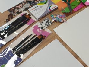 Zdjęcie przedstawiające drewniany stół, na którym rozłożone są materiały plastyczne i kartki papieru. Widzimy tu pędzle malarskie, pudełko z pastelami olejnymi marki Pentel, małe nożyczki, kolorowe druciki, woreczek z cekinami oraz ruchome plastikowe oczka do przyklejania. W dolnej części kartka z wydrukowanym wierszem o tytule „*** [Las wchodzi...]" autorstwa Eevy-Liisy Manner. W rogu kadru widać fragment dłoni.