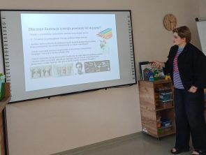 Po prawej stronie stoi kobieta (prowadząca). W prawej ręce trzyma wskaźnik, którym wskazuje na duży ekran interaktywny zawieszony na ścianie. Na ekranie wyświetlony jest slajd z pytaniem: „Dlaczego ilustracja i poezja powinny iść w parze?"