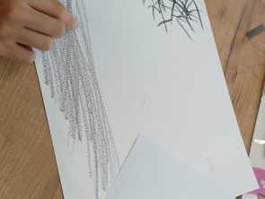 Zdjęcie przedstawia zbliżenie na dłonie osoby, która rysuje na białej kartce leżącej na drewnianym stole. Uczestnik warsztatów używa czarnego pastelu, tworząc rysunek przedstawiający drzewo. W dolnej części kadru na stole leży mniejsza kartka z wydrukowanym wierszem Haliny Poświatowskiej pt. „Na krawędzi mijania". W tle widoczne są inne drobne przybory, takie jak naklejki czy kolorowe patyczki.