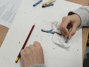 Na zdjęciu widoczne są dłonie uczestnika warsztatów – jedna ręka przytrzymuje kartkę, a druga rysuje szarym pastelem. Na środku papieru widać powstający szkic. Wokół na stole leżą kredki pastelowe, czerwony ołówek, klej w płynie, nożyczki, taśma oraz kartka z tekstem wiersza.