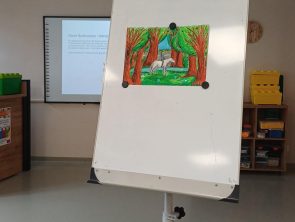 Zdjęcie przedstawia flipchart. Do tablicy za pomocą magnesów przyczepiono kolorowy rysunek przedstawiający białego konia z jeźdźcem, idącego przez gęsty las.