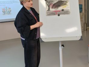 Zdjęcie przedstawia prowadzącą warsztaty kobietę stojącą obok tablicy flipchart na której wisi przypięty magnesami rysunek.