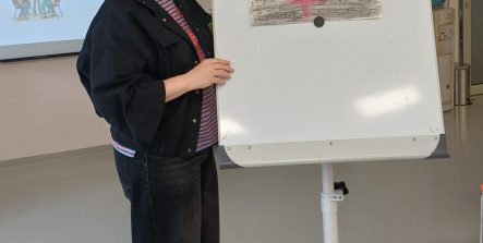 Zdjęcie przedstawia prowadzącą warsztaty kobietę stojącą obok tablicy flipchart na której wisi przypięty magnesami rysunek.