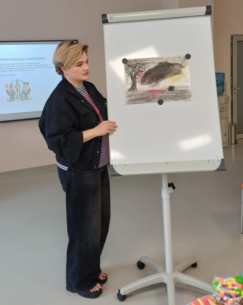 Zdjęcie przedstawia prowadzącą warsztaty kobietę stojącą obok tablicy flipchart na której wisi przypięty magnesami rysunek.