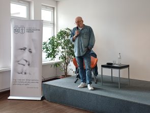Mężczyzna stojący na niewielkiej scenie przemawia do publiczności, trzymając mikrofon w dłoni. Obok niego znajduje się roll-up Miejskiej Biblioteki Publicznej w Zielonce Woli z czarno-białym portretem i cytatem. W tle widać duże okna, roślinę doniczkową, krzesło oraz stolik z butelką wody i szklanką. Wnętrze jest jasne i minimalistyczne.