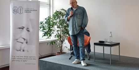 Mężczyzna stojący na niewielkiej scenie przemawia do publiczności, trzymając mikrofon w dłoni. Obok niego znajduje się roll-up Miejskiej Biblioteki Publicznej w Zielonce Woli z czarno-białym portretem i cytatem. W tle widać duże okna, roślinę doniczkową, krzesło oraz stolik z butelką wody i szklanką. Wnętrze jest jasne i minimalistyczne.