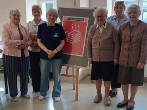 Na zdjęciu widać grupę sześciu kobiet. W centrum znajduje się sztaluga z czerwonym plakatem promującym Dyskusyjny Klub Książki (DKK). Na plakacie widnieje grafika stosu książek oraz napis „Dobre książki i kawa".