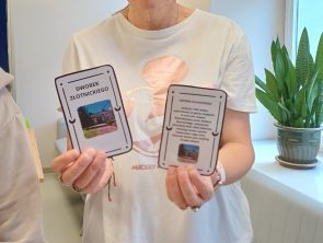Kobieta w okularach i białej koszulce stoi i trzyma w dłoniach dwie białe karty z napisami „Dworek Złotnickiego" oraz zdjęciami budynku i krótkim opisem.