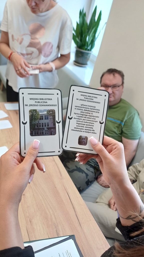 Zdjęcie na którym na pierwszym planie są dwie dłonie trzymające karty z napisami „Miejska Biblioteka Publiczna" i zdjęciami budynku oraz krótkim opisem. W tle widać siedzące przy stole osoby.
