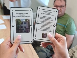 Zdjęcie na którym na pierwszym planie są dwie dłonie trzymające karty z napisami „Miejska Biblioteka Publiczna" i zdjęciami budynku oraz krótkim opisem. W tle widać siedzące przy stole osoby.
