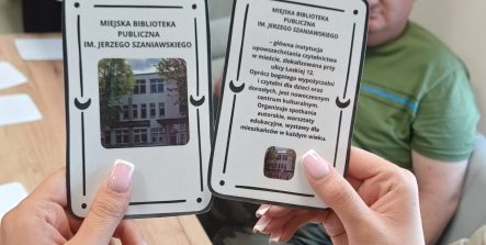 Zdjęcie na którym na pierwszym planie są dwie dłonie trzymające karty z napisami „Miejska Biblioteka Publiczna" i zdjęciami budynku oraz krótkim opisem. W tle widać siedzące przy stole osoby.