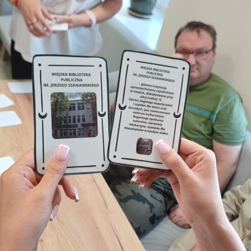 Zdjęcie na którym na pierwszym planie są dwie dłonie trzymające karty z napisami „Miejska Biblioteka Publiczna" i zdjęciami budynku oraz krótkim opisem. W tle widać siedzące przy stole osoby.