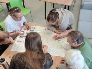 Grupa osób siedzi wokół drewnianego stołu, na którym rozłożona jest duża mapa miasta z zaznaczonymi punktami.
