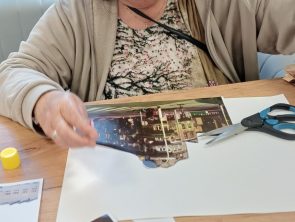 Kobieta w szarej rozpinanej bluzie siedzi przy stole, tworząc kolaż, obok leżą nożyczki.
