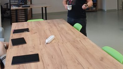 Zdjęcie przedstawia mężczyznę prowadzącego wykład. Stoi on za dużym, drewnianym stołem, trzymając w ręku filiżankę. W tle widać rzutnik wyświetlający grafikę z pytaniem "KIM BYŁ BOLESŁAW LEŚMIAN?"