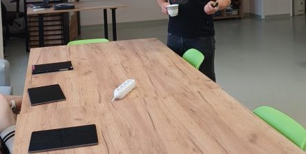 Zdjęcie przedstawia mężczyznę prowadzącego wykład. Stoi on za dużym, drewnianym stołem, trzymając w ręku filiżankę. W tle widać rzutnik wyświetlający grafikę z pytaniem "KIM BYŁ BOLESŁAW LEŚMIAN?"