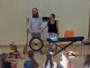 Dwoje instruktorów stoi obok dużego gongu i keyboardu, a grupa dzieci siedzi przed nimi na kolorowych poduszkach, słuchając.