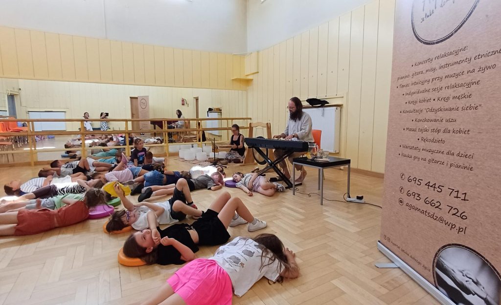 Grupa dzieci leży na podłodze w sali podczas koncertu relaksacyjnego. Mężczyzna w tle gra na pianinie cyfrowym, a obok niego widoczne są misy dźwiękowe i gong. Po prawej stronie stoi baner reklamowy.