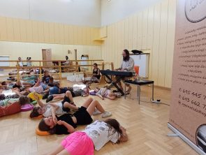 Grupa dzieci leży na podłodze w sali podczas koncertu relaksacyjnego. Mężczyzna w tle gra na pianinie cyfrowym, a obok niego widoczne są misy dźwiękowe i gong. Po prawej stronie stoi baner reklamowy.