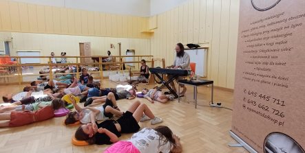 Grupa dzieci leży na podłodze w sali podczas koncertu relaksacyjnego. Mężczyzna w tle gra na pianinie cyfrowym, a obok niego widoczne są misy dźwiękowe i gong. Po prawej stronie stoi baner reklamowy.