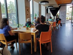 Sala kawiarni literackiej z dużymi oknami i stolikami, przy których siedzą uczestnicy spotkania autorskiego. Na końcu sali widoczna jest osoba prowadząca wydarzenie.