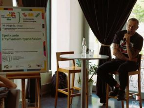 Mężczyzna siedzi przy wysokim stoliku i mówi do mikrofonu podczas spotkania autorskiego. Obok znajduje się plansza informacyjna zapowiadająca spotkanie z Tymonem Tymańskim w ramach Dni Zduńskiej Woli.