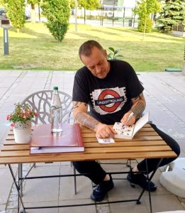 Tymon Tymański siedzi przy stoliku na zewnątrz kawiarni literackiej i podpisuje książkę. Na stole stoją butelka wody, szklanka oraz kronika, a w tle widać zieleń i otwartą przestrzeń.