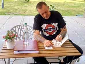 Tymon Tymański siedzi przy stoliku na zewnątrz kawiarni literackiej i podpisuje książkę. Na stole stoją butelka wody, szklanka oraz kronika, a w tle widać zieleń i otwartą przestrzeń.