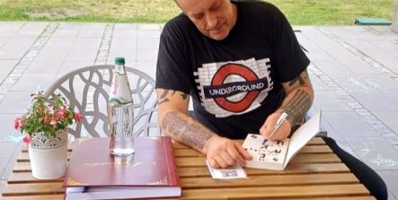Tymon Tymański siedzi przy stoliku na zewnątrz kawiarni literackiej i podpisuje książkę. Na stole stoją butelka wody, szklanka oraz kronika, a w tle widać zieleń i otwartą przestrzeń.