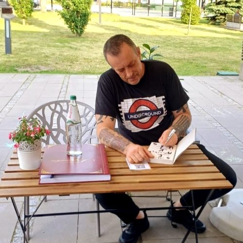 Tymon Tymański siedzi przy stoliku na zewnątrz kawiarni literackiej i podpisuje książkę. Na stole stoją butelka wody, szklanka oraz kronika, a w tle widać zieleń i otwartą przestrzeń.