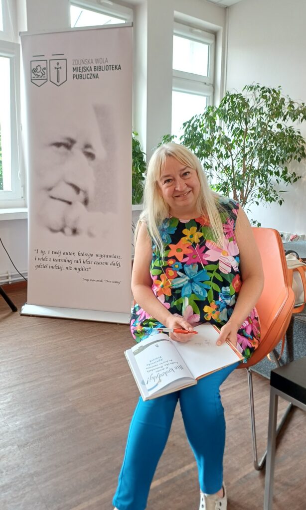 Kobieta siedzi na pomarańczowym krześle i trzyma na kolanach otwartą książkę dla dzieci pod tytułem „Nie krokodyluj!" oraz czerwony długopis. Po jej lewej stronie stoi biały baner Miejskiej Biblioteki Publicznej w Zduńskiej Woli.