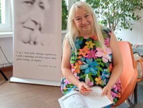 Kobieta siedzi na pomarańczowym krześle i trzyma na kolanach otwartą książkę dla dzieci pod tytułem „Nie krokodyluj!" oraz czerwony długopis. Po jej lewej stronie stoi biały baner Miejskiej Biblioteki Publicznej w Zduńskiej Woli.