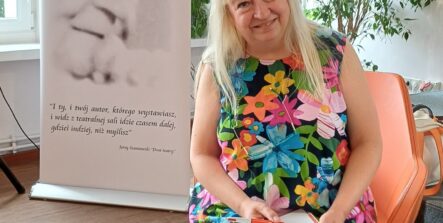 Kobieta siedzi na pomarańczowym krześle i trzyma na kolanach otwartą książkę dla dzieci pod tytułem „Nie krokodyluj!" oraz czerwony długopis. Po jej lewej stronie stoi biały baner Miejskiej Biblioteki Publicznej w Zduńskiej Woli.