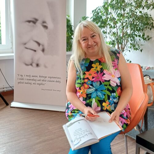 Kobieta siedzi na pomarańczowym krześle i trzyma na kolanach otwartą książkę dla dzieci pod tytułem „Nie krokodyluj!" oraz czerwony długopis. Po jej lewej stronie stoi biały baner Miejskiej Biblioteki Publicznej w Zduńskiej Woli.