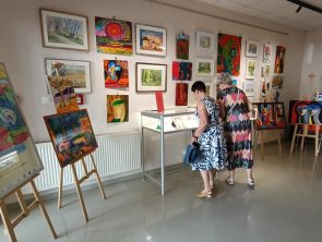 Dwie kobiety oglądają wystawę obrazów w galerii biblioteki, stojąc tyłem do obiektywu przy szklanej gablocie. Po lewej stronie widać plakat wernisażu, a w głębi sali na sztalugach i ścianach wiszą kolorowe dzieła sztuki.