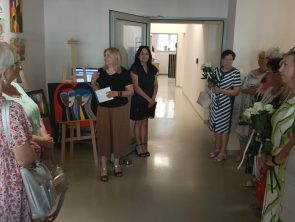 Grupa osób zebrana w korytarzu biblioteki podczas otwarcia wystawy. Dwie kobiety na środku witają gości, podczas gdy pozostali , z których część trzyma białe róże, słuchają. W tle widoczne są sztalugi z obrazami.