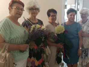 Półprofil pięciu kobiet stojących obok siebie i pozujących do zdjęcia w galerii. Trzy z nich trzymają w dłoniach bukiety kwiatów,