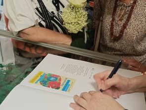 Zbliżenie na dłonie osoby piszącej w pamiątkowej księdze wyłożonej na szklanej gablocie.
