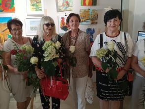Sześć kobiet stoi w rzędzie na tle ściany z obrazami, pozując wspólnie do zdjęcia. Każda z nich trzyma w rękach białą różę lub bukiet jasnych kwiatów. Wszystkie patrzą w stronę aparatu.