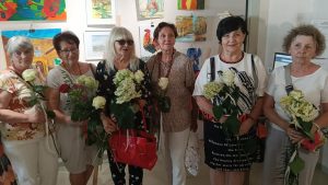 Sześć kobiet stoi w rzędzie na tle ściany z obrazami, pozując wspólnie do zdjęcia. Każda z nich trzyma w rękach białą różę lub bukiet jasnych kwiatów. Wszystkie patrzą w stronę aparatu.