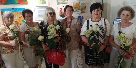 Sześć kobiet stoi w rzędzie na tle ściany z obrazami, pozując wspólnie do zdjęcia. Każda z nich trzyma w rękach białą różę lub bukiet jasnych kwiatów. Wszystkie patrzą w stronę aparatu.
