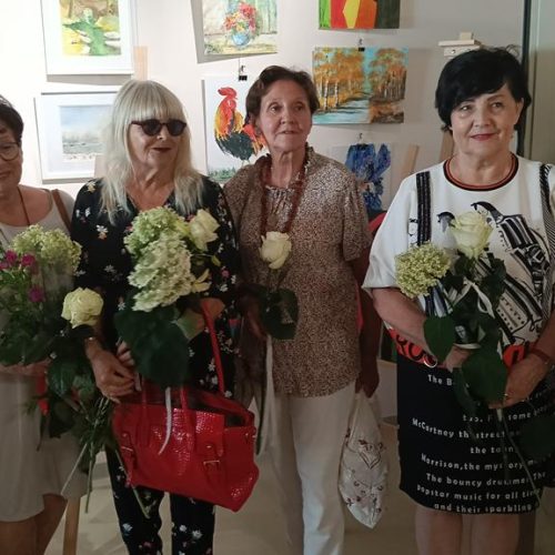 Sześć kobiet stoi w rzędzie na tle ściany z obrazami, pozując wspólnie do zdjęcia. Każda z nich trzyma w rękach białą różę lub bukiet jasnych kwiatów. Wszystkie patrzą w stronę aparatu.