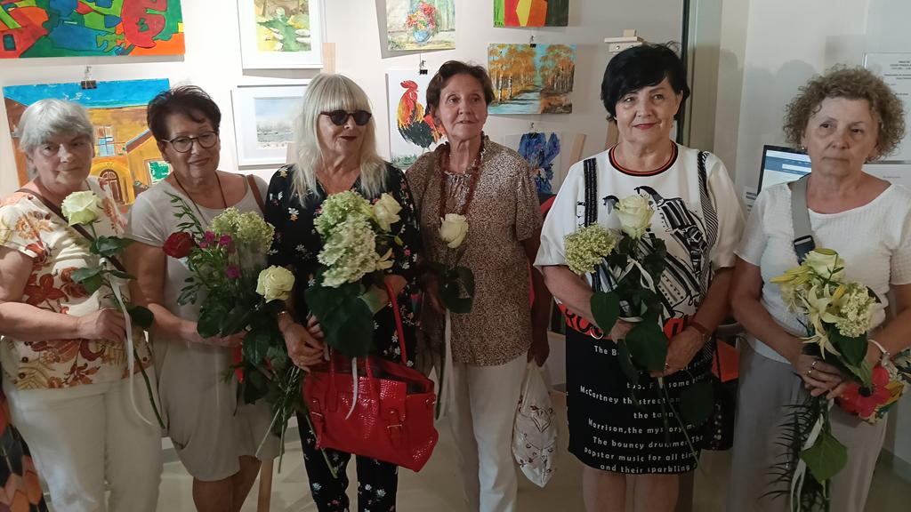 Sześć kobiet stoi w rzędzie na tle ściany z obrazami, pozując wspólnie do zdjęcia. Każda z nich trzyma w rękach białą różę lub bukiet jasnych kwiatów. Wszystkie patrzą w stronę aparatu.